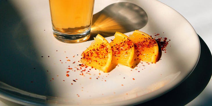 Vaso servido con tequila encima de un plato con rebanadas de naranja espolvoreadas con chile en polvo sobre una mesa blanca y un fondo negro en un negocio de tequila.