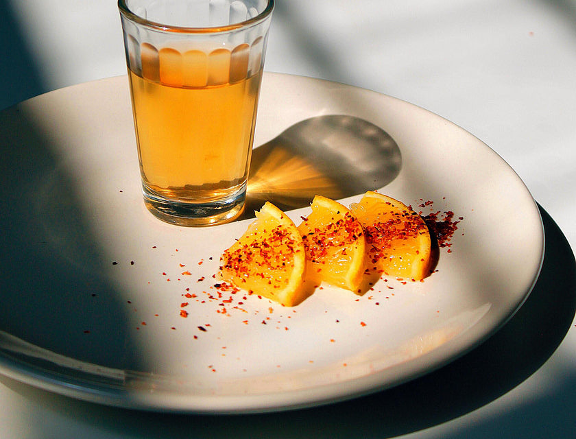 Vaso servido con tequila encima de un plato con rebanadas de naranja espolvoreadas con chile en polvo sobre una mesa blanca y un fondo negro en un negocio de tequila.