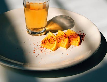 Vaso servido con tequila encima de un plato con rebanadas de naranja espolvoreadas con chile en polvo sobre una mesa blanca y un fondo negro en un negocio de tequila.