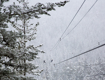 Dos personas suspendidas en una tirolesa con un fondo de pinos nevados en un negocio de tirolesa.