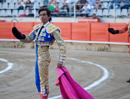Dos toreros en el ruedo vistiendo el traje de luces color azul y color rojo respectivamente con público al fondo.