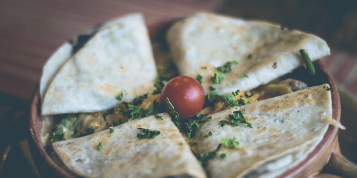 Una plato con cuatro quesadillas encima de una cama de queso con cilantro y un jitomate cherry en un puesto de quesadillas.