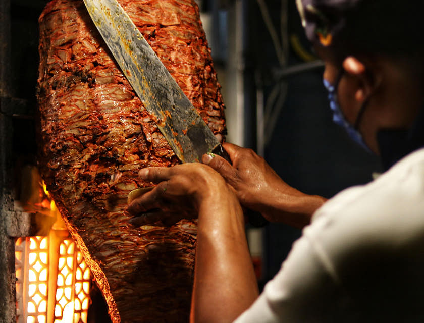 Una pieza de carne al pastor y un taquero frente a ella preparando una orden de tacos al pastor en un puesto de tacos al pastor.