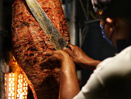 Una pieza de carne al pastor y un taquero frente a ella preparando una orden de tacos al pastor en un puesto de tacos al pastor.