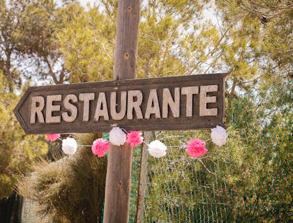 Señalamiento de un restaurante campestre colgado de un poste de madera entre árboles y flores de papel.