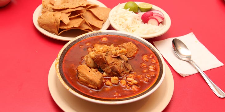 Platón de pozole sobre una mesa con mantel rojo, con plato de totopos, cebolla, rábano, limón y una cuchara al lado en un restaurante de comida jalisciense.