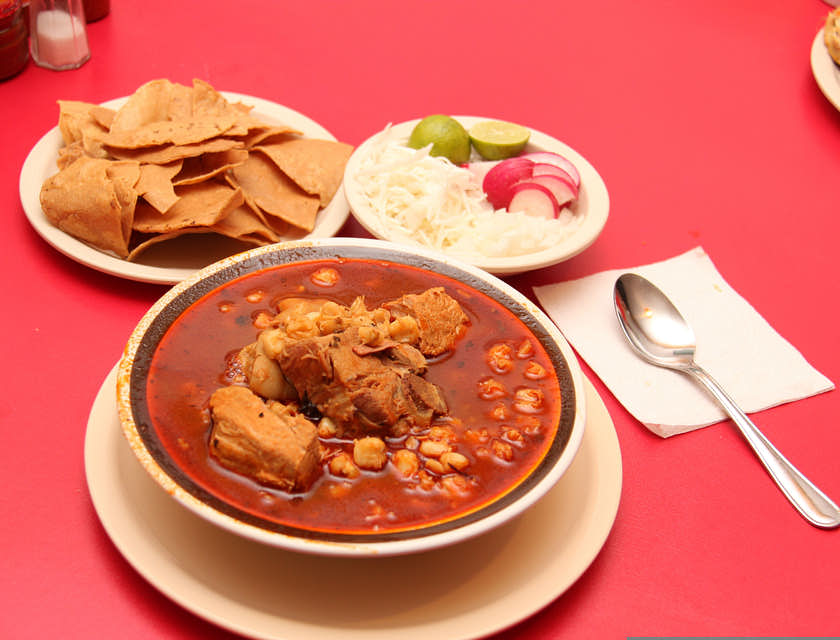 Platón de pozole sobre una mesa con mantel rojo, con plato de totopos, cebolla, rábano, limón y una cuchara al lado en un restaurante de comida jalisciense.