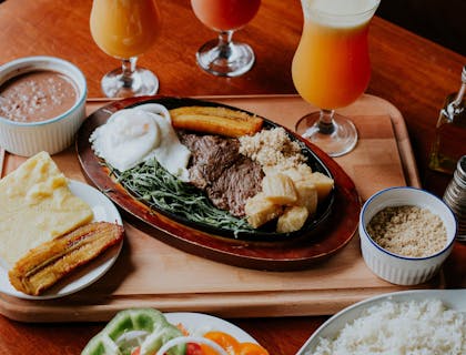 Tres vasos de jugos a un lado de un platillo de carne, verduras y otro con un plátano y pan en un restaurante de minutas.