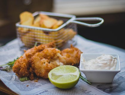 Pescado frito sobre un periódico con medio limón, aderezo y una canastilla de papas al aceite en un restaurante inglés.