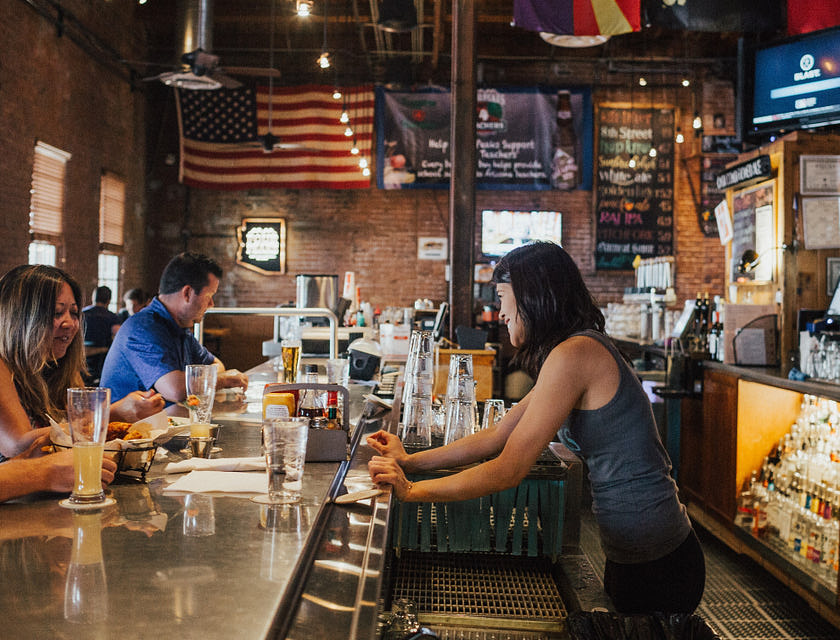 Una mujer atendiendo a dos clientes en la barra de un restobar.