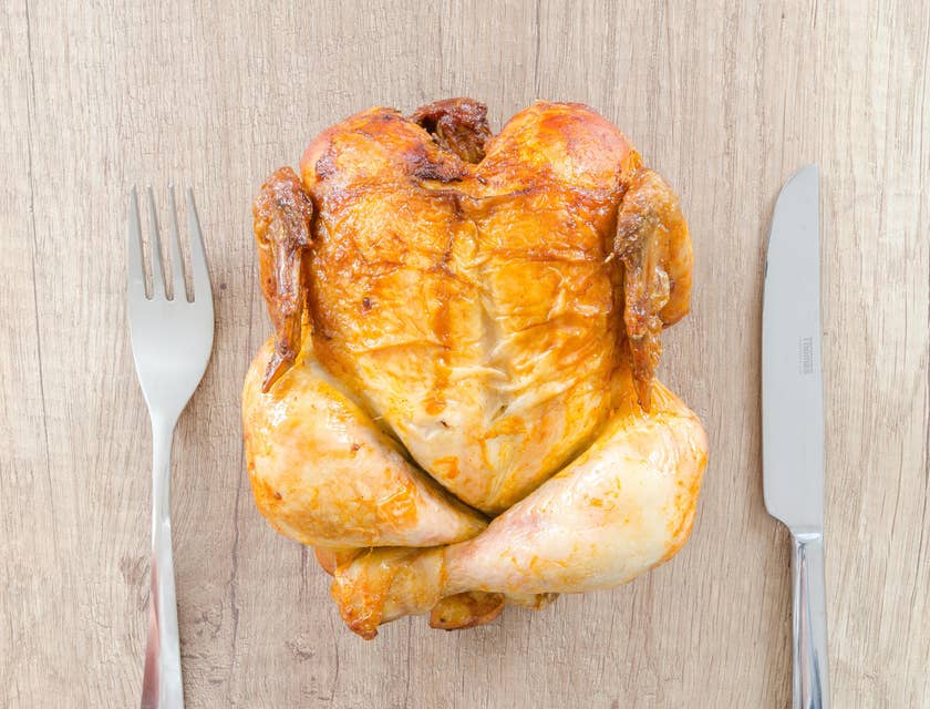Un pollo rostizado en una mesa de madera con dos cubiertos a un lado en una rosticería.