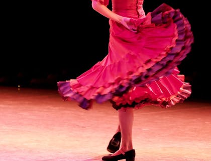 Una mujer de espaldas bailando flamenco vistiendo ropa flamenca con un fondo negro sobre duela de madera.