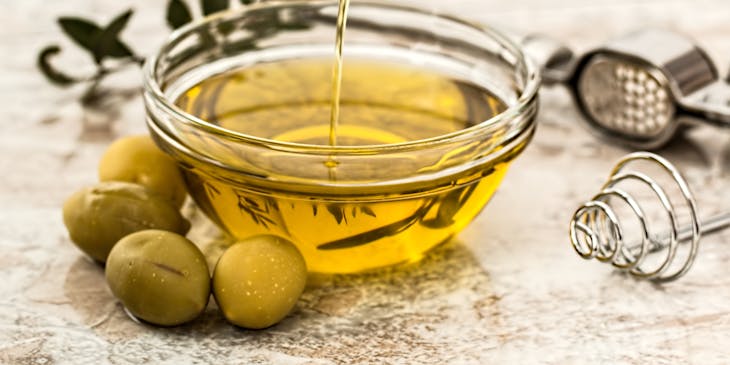 Olive oil poured into a glass container with green olives arranged around the container.