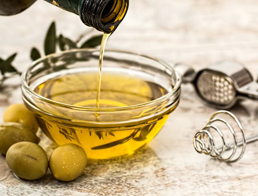 Olive oil poured into a glass container with green olives arranged around the container.