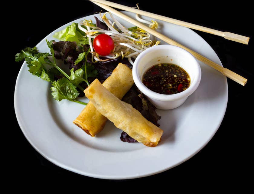 Ein Teller mit Frühlingsrollen, Sojasauce, frischem Gemüse und Essstäbchen liegt in einem pan-asiatischen Restaurant auf einem dunklen Tisch.