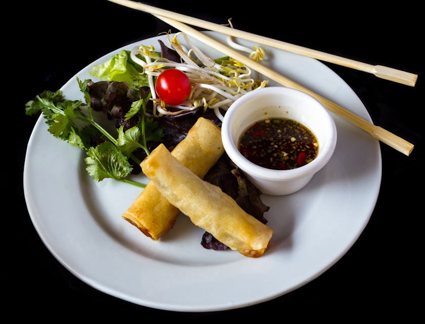 Ein Teller mit Frühlingsrollen, Sojasauce, frischem Gemüse und Essstäbchen liegt in einem asiatischen Restaurant auf einem dunklen Tisch.