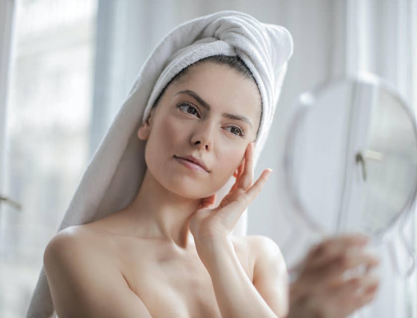 Una donna con i capelli avvolti in un asciugamano che si tocca il viso e si guarda allo specchio.