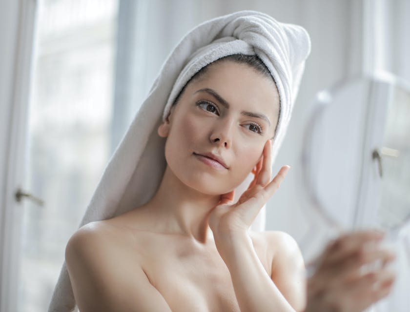 Una donna con i capelli avvolti in un asciugamano che si tocca il viso e si guarda allo specchio.