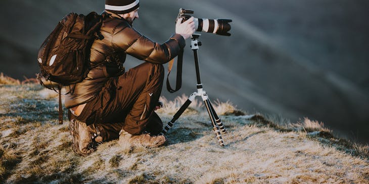 A photographer crouched on a mountaintop taking pictures for his photography business.