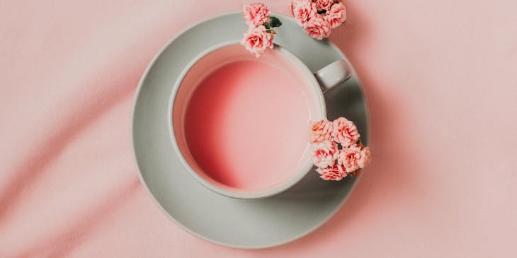 A cup of pink tea surrounded by flowers displayed against a pink background.