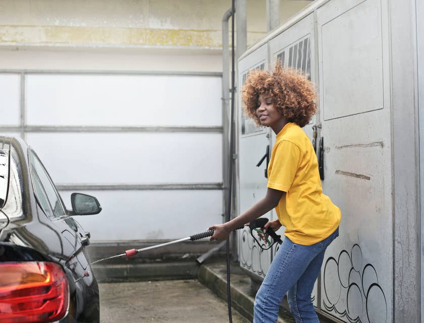 Mulher usando um uniforme amarelo e realizando um serviço de lavagem de alta pressão em um carro preto.