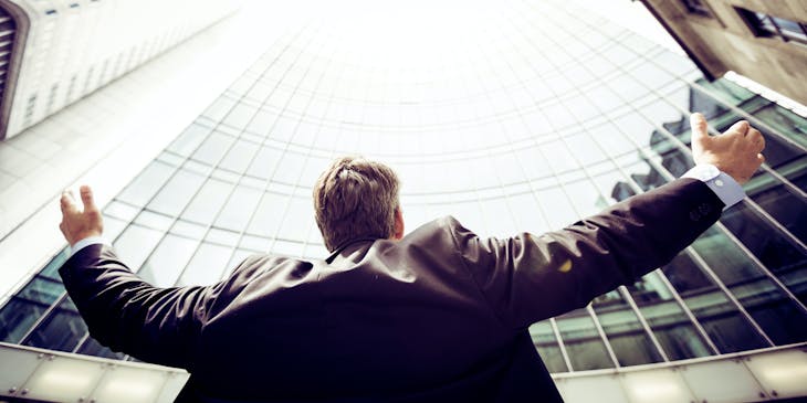 Man standing at the base of tall, powerful buildings raising his arms