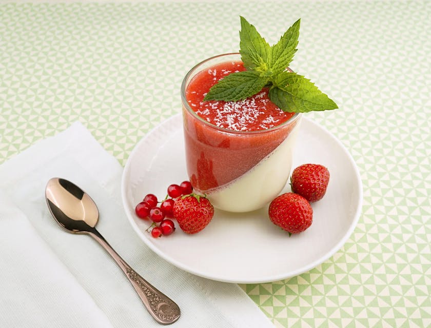 Un pouding aux fraises et à la vanille présenté avec une cuillère sur une table blanche.