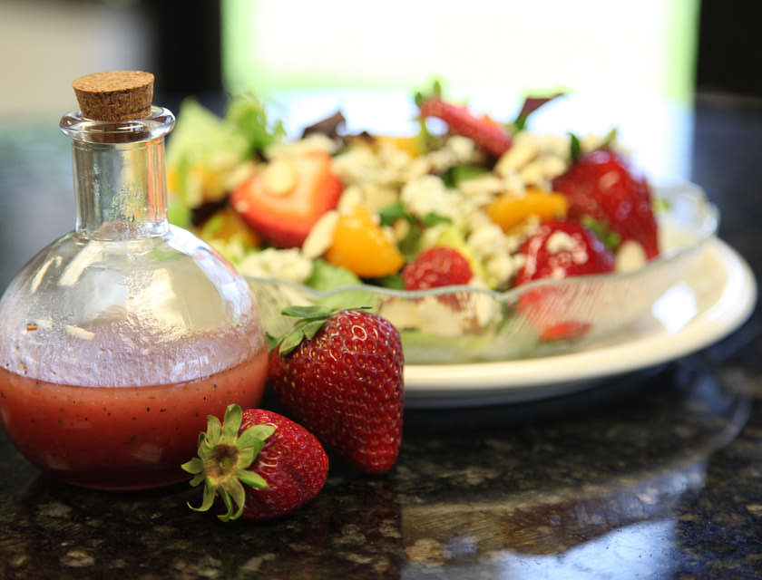 Aderezo de fresa en una botella de vidrio a un lado de una ensalada de lechuga con frutas en un negocio de aderezos de ensaladas.