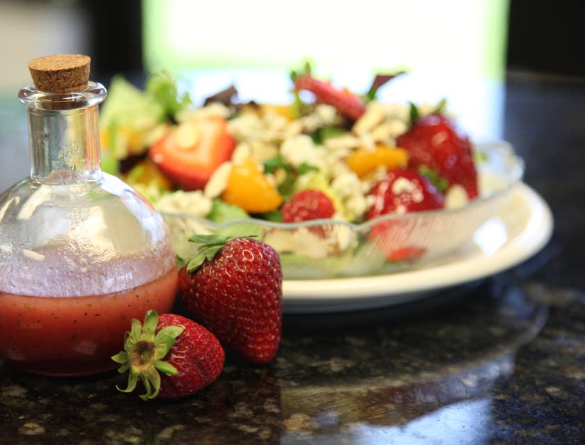 Aderezo de fresa en una botella de vidrio a un lado de una ensalada de lechuga con frutas en un negocio de aderezos de ensaladas.