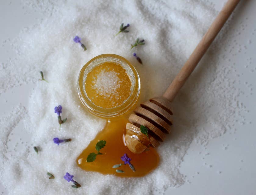 Ein Honig-Körperpeeling in einem Glasgefäß liegt auf einer mit Salz und Blüten bestreuten Oberfläche.