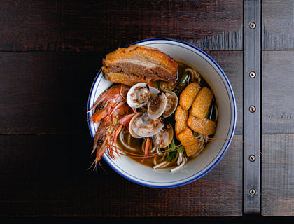 White bowl of noodles in broth with a variety of seafood on a dark wooden table at a Singaporean restaurant.