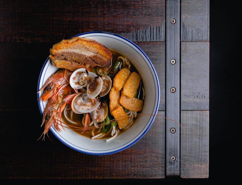 White bowl of noodles in broth with a variety of seafood on a dark wooden table at a Singaporean restaurant.