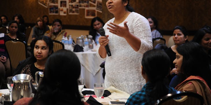 A woman addressing multiple tables of people during speech training.