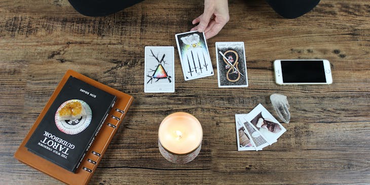 A hand touching a tarot card next to books, a candle, photos, a crystal, and a phone.