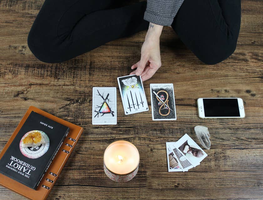 A hand touching a tarot card next to books, a candle, photos, a crystal, and a phone.
