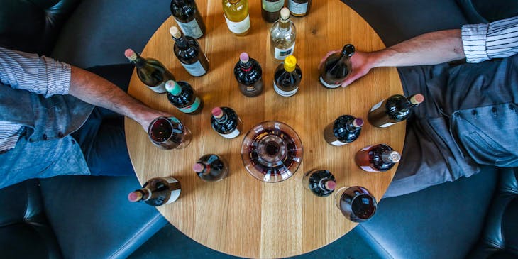 Two men sitting at a round wooden table holding many wine bottles, a wine glass, and a glass decanter in a tasting class.