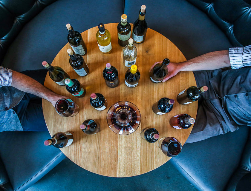 Dos hombres sentados en una mesa redonda de madera sosteniendo muchas botellas de vino, una copa de vino y una jarra de vidrio en una escuela de cata.