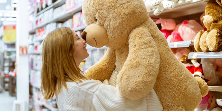 Woman in a toy store taking a big brown teddy bear down from a shelf with more rows of teddy bears in the background.