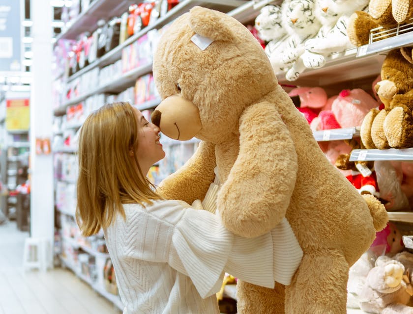 Woman in a toy store taking a big brown teddy bear down from a shelf with more rows of teddy bears in the background.