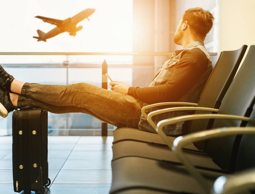Homem sentado na cadeira de um aeroporto e olhando pela janela enquanto um avião decola.