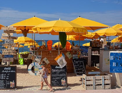 Ein kleines Mädchen im Badeanzug vor einer tropischen Strandbar mit gelben Sonnenschirmen und Schiefer-Werbetafeln.