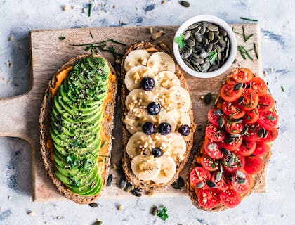Drei, mit veganen Aufstrichen belegte Brote liegen neben einer Schale mit Kürbiskernen auf einem Holzschneidebrett.