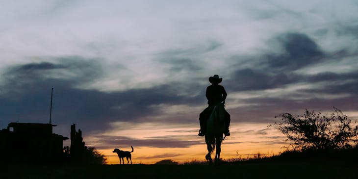 Silhueta de um cowboy em cima de um cavalo contra o pôr do sol no faroeste.
