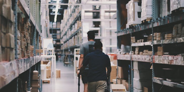 Two men walking down an aisle at a wholesale store.