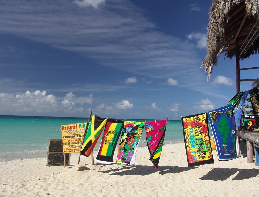 Una playa con un tendedero con toallas con temáticas jamaiquinas a lado de una palapa de un negocio con nombre jamaiquino.