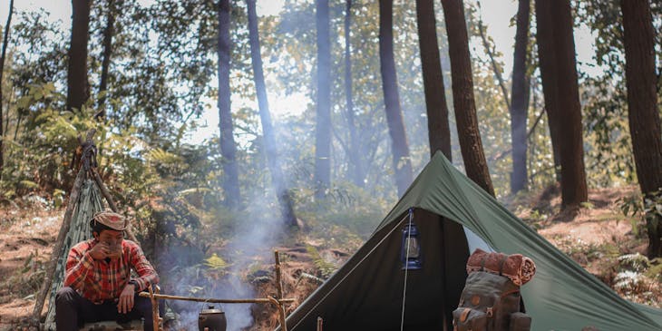 Un campista tomando café a un lado de su casa de campaña en un campamento en el bosque.