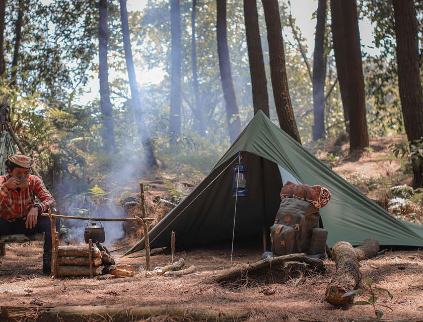 Un campista tomando café a un lado de su casa de campaña en un campamento en el bosque.