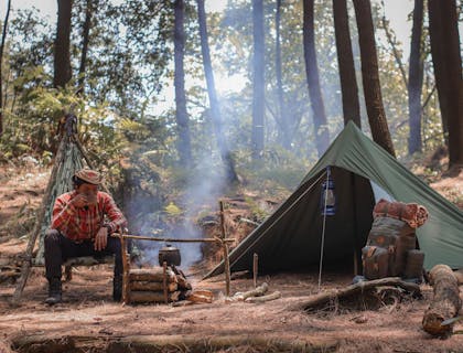 Un campista tomando café a un lado de su casa de campaña en un campamento en el bosque.