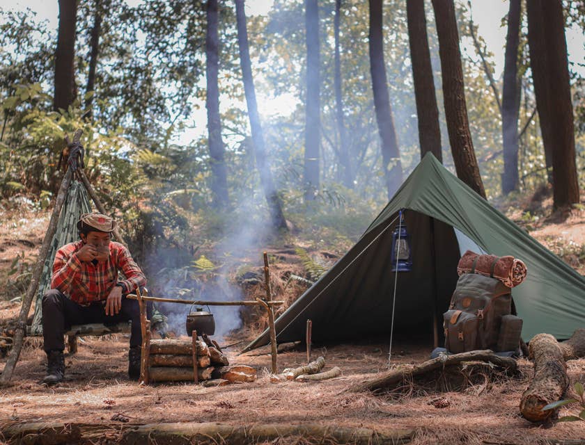 Un campista tomando café a un lado de su casa de campaña en un campamento en el bosque.