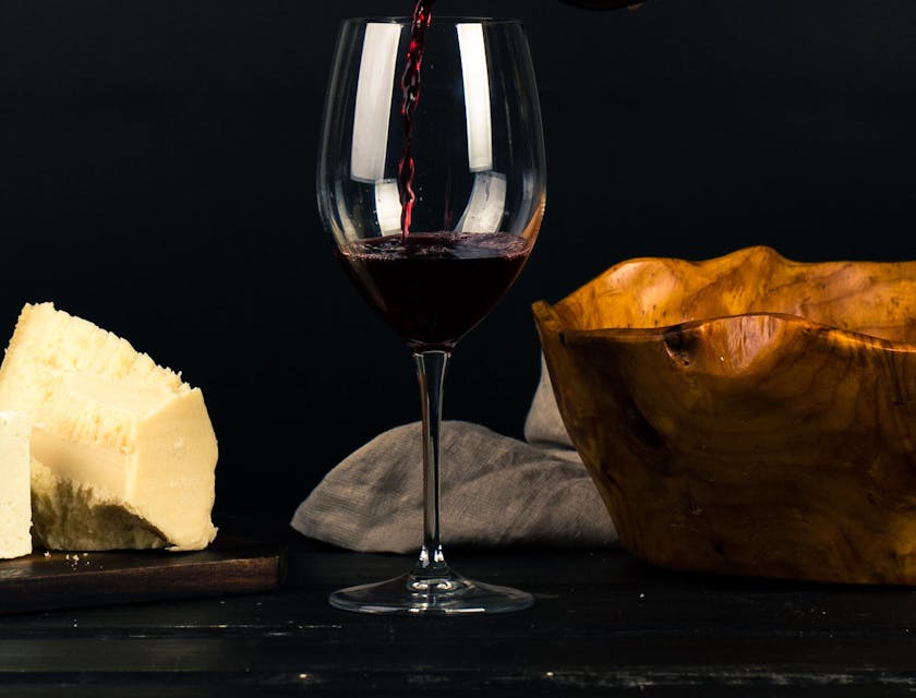 Una persona sirviendo una copa de vino tinto sobre una mesa con un plato de quesos en un bar de vino.